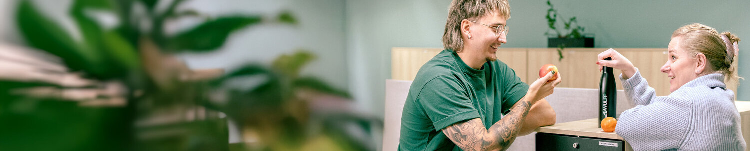 Kaksi nuorta toimistolla, toislla juomapullo ja toisella omena.