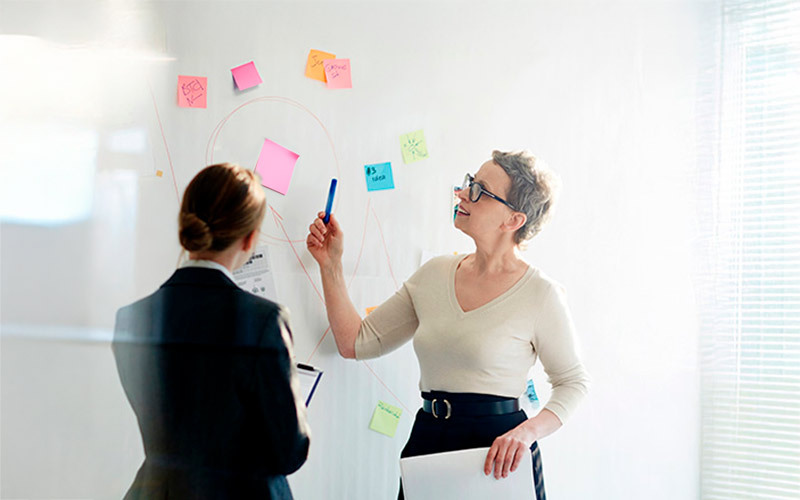 Kaksi henkilöä valkotaulun edessä, jossa paljon värikkäitä viestilappuja.