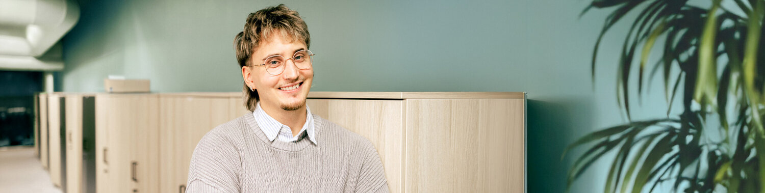 Nuorehko silmälaispäinen miesoletettu hymyilee toimistolla kädet puuskassa